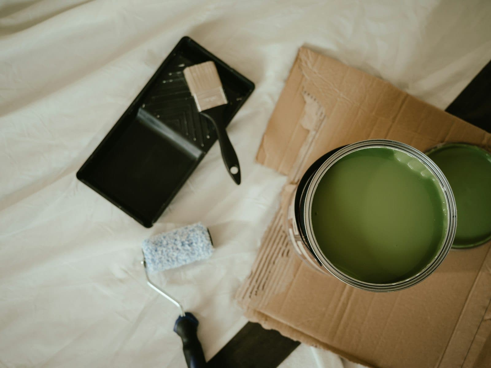 Flat lay of an open sage green paint can with tray, brush, and mini roller on a drop cloth
