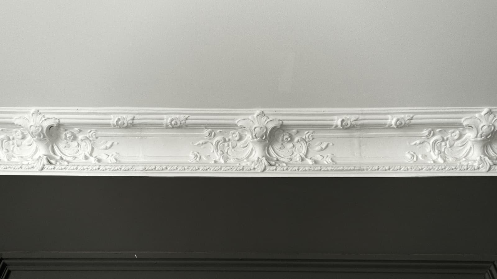 Ornate white crown molding running along a ceiling line above a dark-painted wall, shown in close-up